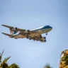 Side view of the iconic blue and white Boeing Air Force One jet landing under a clear blue sky with palm trees in the foreground.