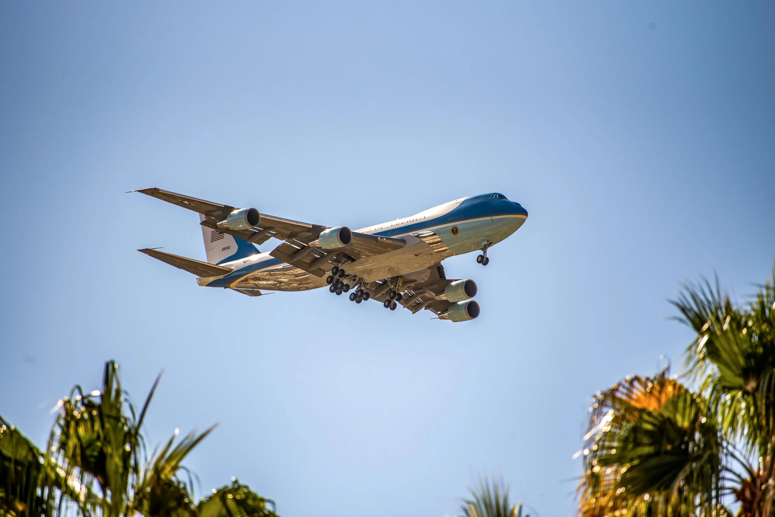 Side view of the iconic blue and white Boeing Air Force One jet landing under a clear blue sky with palm trees in the foreground.
